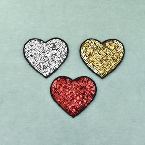 Peut inclure: Trois écussons en forme de cœur recouverts d'une couche dense de petites paillettes réfléchissantes. Un écusson est argenté, un est doré et un est rouge. Chaque cœur est souligné par une fine bordure noire, créant un contraste marqué.