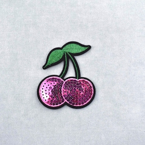 Peut inclure: Écusson décoratif représentant deux cerises roses à paillettes, bordées de noir. Les cerises sont reliées par une tige noire avec deux feuilles vertes. Fond gris clair.