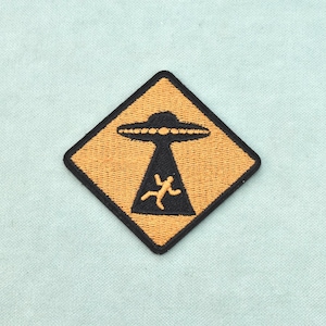 May include: A diamond-shaped embroidered patch with a black border on an orange background. The design features a black UFO hovering above a light orange human figure, with a black beam of light extending downward from the craft to the figure.