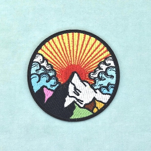 Könnte beinhalten: Ein kreisförmiger bestickter Aufnäher mit einer Berglandschaft, einer roten Sonne und gelben Sonnenstrahlen. Das Design zeigt stilisierte hellblaue Wolken auf beiden Seiten eines dunklen Berggipfels mit weißem Schnee und einer grünen Basis.