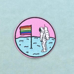 Op de afbeelding: Een ronde geborduurde patch met een witte astronaut die op een lichtblauw maanoppervlak staat. Naast de astronaut staat een vlaggenmast met een regenboogkleurige vlag. De achtergrond van de scène is een effen lichtroze cirkel.