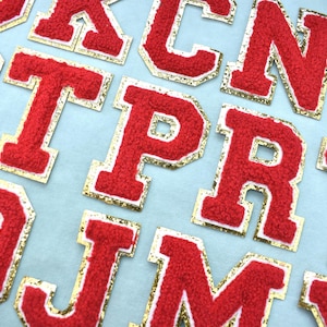 Op de afbeelding: Close-up van rode vilt varsity letters met gouden rand. De letters zijn in verschillende lettertypen en maten, gerangschikt op een lichtblauwe achtergrond. Ideaal voor knutselprojecten, deze letters geven kleding en accessoires een klassieke uitstraling.