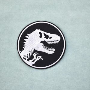 Peut inclure: Un écusson rond noir avec un crâne de dinosaure blanc brodé, encadré d'un cercle blanc. Le crâne de dinosaure présente des dents et une structure osseuse détaillées. L'écusson est posé sur un fond bleu-vert clair.