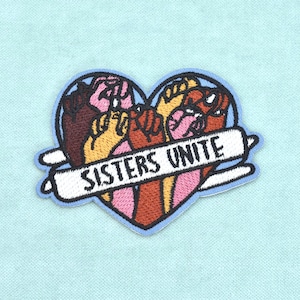 Può includere: Una toppa ricamata a forma di cuore con un bordo azzurro. Il design presenta diversi pugni alzati in varie tonalità di pelle, tra cui marrone, beige e rosa. Un nastro bianco al centro contiene il testo nero SISTERS UNITE.