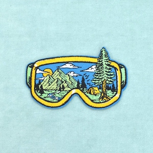 Può includere: Una toppa ricamata a forma di maschera da sci con montatura gialla. L'area della lente presenta una scena all'aperto dettagliata con montagne verdi, un fiume blu, un sole giallo, nuvole bianche, un pino alto e una piccola tenda arancione.