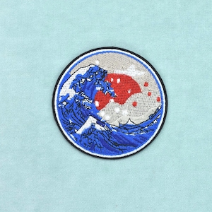 Pode incluir: Um emblema bordado circular apresentando um design estilizado de onda oceânica azul com cristas de espuma branca. Atrás da onda, há um grande sol circular vermelho sobre um fundo cinza claro. O design é contornado por uma borda preta.