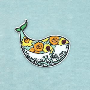 Peut inclure: Un écusson brodé en forme de baleine avec une queue vert clair. La partie supérieure présente un motif de tournesols jaunes et orange aux centres sombres, tandis que la section inférieure affiche des vagues stylisées blanches et vert clair.
