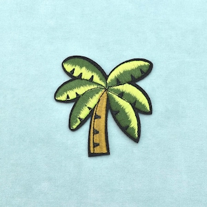 Pode incluir: Um emblema bordado em forma de palmeira com tronco castanho e folhas verdes. O design apresenta um contorno preto bordado que delineia a árvore, com fio verde claro e escuro usado para criar textura e profundidade nas folhas da palmeira.