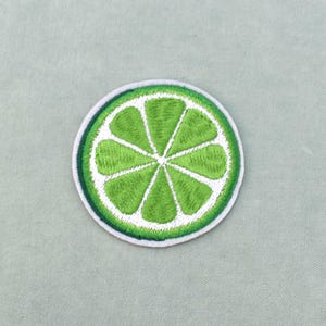 Peut inclure: Écusson brodé représentant une tranche de citron vert. Le patch a un centre vert vif avec des segments blancs, une bordure blanche et un contour vert. Le patch est circulaire et posé sur un fond en tissu vert clair.