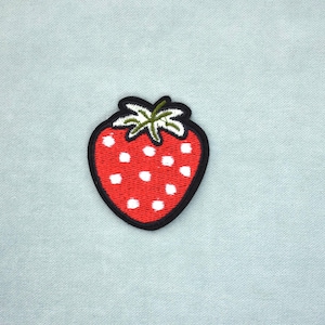 Peut inclure: Écusson brodé en forme de fraise, rouge avec des graines blanches, une tige et des feuilles vertes. Le patch a un bord noir et est posé sur un fond bleu clair. Idéal pour les vêtements ou accessoires.