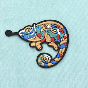 Puede incluir: Un parche bordado termoadhesivo que muestra un camaleón con la cola enroscada. El diseño utiliza un patrón tipo mosaico con hilos de colores beige, azul, rojo y blanco, delineado con hilo negro, representando al lagarto con la lengua extendida.