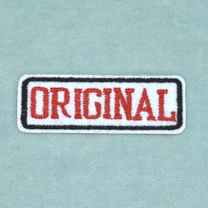 Op de afbeelding: Een rechthoekige geborduurde patch met het woord ORIGINAL in dikke rode hoofdletters. De tekst is gecentreerd op een witte achtergrond, omringd door een dikke zwarte rand en afgewerkt met een witte buitenrand op lichtblauwe stof.