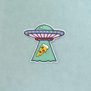 Könnte beinhalten: Ein gestickter Aufnäher, der ein hellgrünes UFO mit violettem Rand und gelben Sternen zeigt. Das Raumschiff hat eine rosafarbene Unterseite und sendet einen hellgrünen Traktorstrahl aus, der ein Stück Peperoni-Pizza mit gelbem Käse anhebt.