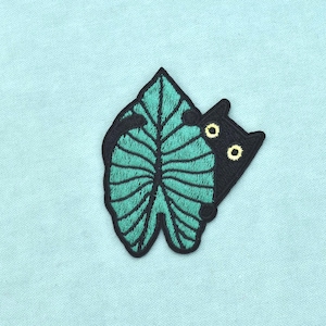 Op de afbeelding: Een geborduurde patch met een zwarte kat die achter een groot, getextureerd blauwgroen blad vandaan kijkt. De kat heeft twee zichtbare gele ogen en zwarte oren, terwijl het blad gedetailleerde donkergroene stiksels heeft voor de nerven.