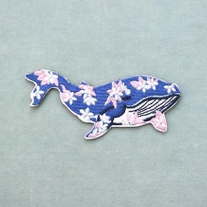 以下が含まれることがあります： 青いクジラの形をした刺繍パッチ。白とピンクの花柄のアクセントが施されています。クジラの腹には白黒の縞模様があります。パッチは水色の背景にあります。