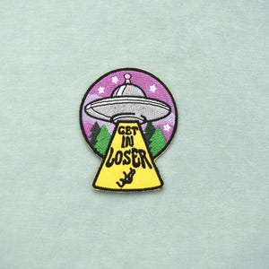 Pode incluir: Patch bordado com um OVNI prateado com o texto "GET IN LOSER" em amarelo. O OVNI está sobre um fundo roxo com estrelas, árvores e uma figura a cair.