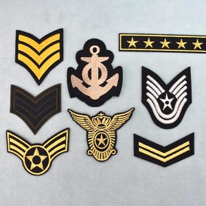Könnte beinhalten: Eine Sammlung von acht bestickten Aufnähern im Militärstil mit verschiedenen Motiven, darunter Winkel, ein zentraler Anker, ein geflügeltes Emblem mit Stern und ein horizontaler Balken mit fünf Sternen, in Gold, Schwarz, Weiß und Beige.