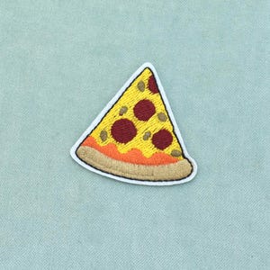Könnte beinhalten: Ein gestickter Aufnäher in Form eines dreieckigen Pizzastücks mit gelbem Käsebelag, dunkelroten Pepperoni-Kreisen, einer orangefarbenen Soßenschicht und einem beigen Krustenrand, alles weiß umrandet auf hellblauem Hintergrund.