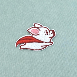 May include: An embroidered patch featuring a white piglet with pink inner ears and a dark red cape, depicted in a flying pose with its arms extended forward. The design has a white border and is rendered in a cartoon style with simple lines.