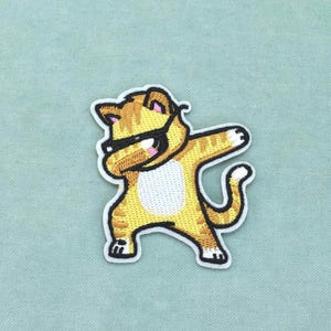 Peut inclure: Un écusson brodé représentant un chat tigré jaune et orange en train de faire le dab. Le chat a un ventre blanc, des contours noirs, un masque noir sur les yeux, une queue courbée et des pattes blanches, avec une bordure blanche.