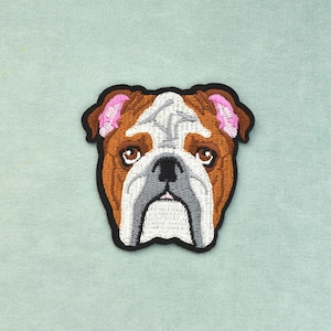 Può includere: Una toppa ricamata raffigurante la testa di un bulldog con pelo marrone e bianco, occhi marrone scuro e orecchie interne rosa. Il disegno è delineato da un bordo nero spesso, con cuciture dettagliate che definiscono i tratti del viso.