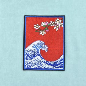 Pode incluir: Um emblema retangular bordado com um fundo vermelho vibrante. O design apresenta uma grande onda azul e branca na parte inferior e um ramo de árvore castanho com flores de cerejeira brancas e cor-de-rosa no canto superior direito.