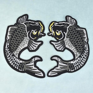 Peut inclure: Deux écussons brodés représentant des poissons koï dans une orientation en miroir. Chaque poisson est réalisé avec des fils noirs, gris et blancs, avec des touches de jaune sur les yeux et la tête, le tout bordé d'un contour noir épais.
