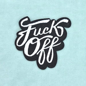 Peut inclure: Un écusson brodé noir présentant le texte blanc "Fuck Off" dans une écriture cursive stylisée. Le lettrage est gras et proéminent, avec la broderie suivant les contours des mots, créant une texture distincte et en relief sur le tissu.