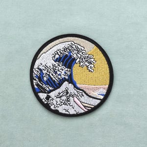 Puede incluir: Parche bordado redondo con un diseño de ola estilizada en tonos azules, blancos y beige, con un sol amarillo y un borde negro. El parche es un artículo decorativo, adecuado para ropa o accesorios.