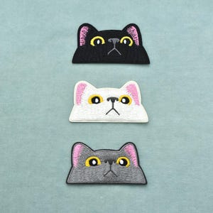 Peut inclure: Trois écussons brodés en forme de tête de chat disposés verticalement. L'écusson du haut est noir, celui du milieu est blanc et celui du bas est gris. Chaque chat a des yeux jaunes, un nez noir, une petite bouche et des oreilles roses.