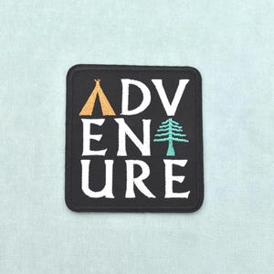 Pode incluir: Um emblema bordado quadrado de cor preta com o texto branco ADVENTURE. A letra A é substituída por um ícone de tipi dourado e a letra T é substituída por um ícone de pinheiro em tom azul-petróleo. O emblema tem uma borda preta sólida.