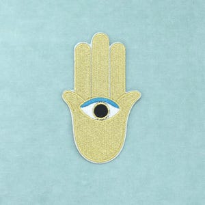 Può includere: Una toppa ricamata color oro a forma di mano di Hamsa, caratterizzata da un motivo a occhio centrale con pupilla nera, iride bianca e dettaglio della palpebra azzurro. La toppa presenta un bordo bianco cucito e una superficie testurizzata.
