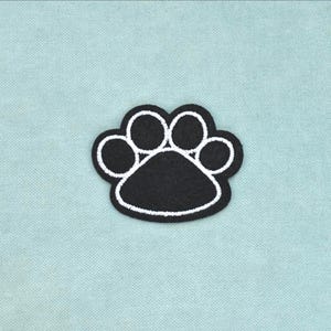 May include: A black embroidered patch in the shape of a paw print. The design features a large central pad and four smaller circular toe pads, all outlined with a thick white stitched border that defines the individual shapes of the paw print.