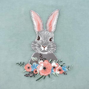 May include: Embroidered image of a grey bunny with pink-lined ears and a floral bouquet. The bunny has detailed whiskers and is surrounded by coral, blue, and white flowers with green leaves, set against a light teal background.