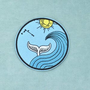 Può includere: Una toppa ricamata circolare con sfondo azzurro e bordo blu scuro. Il design raffigura una coda di balena bianca che emerge da un'onda blu scuro, un sole giallo nella parte superiore centrale e una costellazione di stelle nera.