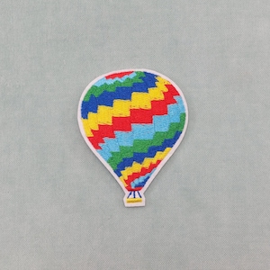 Peut inclure: Écusson brodé d'une montgolfière colorée. Le ballon présente un motif en zigzag rouge, jaune, bleu et vert, souligné de blanc. Le bas du ballon a un petit panier jaune et blanc.