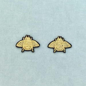 Pode incluir: Dois emblemas bordados com um design de inseto estilizado. Cada emblema apresenta um padrão de fio amarelo dourado formando a silhueta de um besouro com as asas abertas, contornado com fio preto sobre um fundo de tecido azul claro.