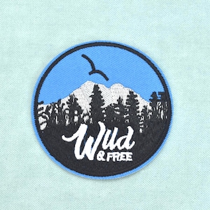 Op de afbeelding: Een ronde geborduurde patch met een lichtblauwe luchtachtergrond, een zwart silhouet van een bos en witte besneeuwde bergen. Een enkele zwarte vogel vliegt boven de bergen. De tekst Wild & Free staat in witte letters op de patch.