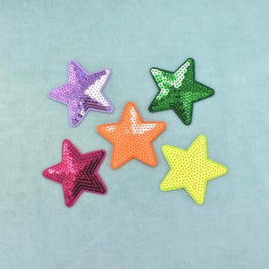 以下が含まれることがあります： 星型のワッペン5つがグループ状に配置されています。紫のスパンコール星、緑のスパンコール星、鮮やかなオレンジのスパンコール星、ホットピンクのスパンコール星、そしてドット状のテクスチャーを持つ黄色の星が含まれています。