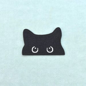 Può includere: Una toppa ricamata nera a forma di testa di gatto con orecchie a punta. La toppa presenta due occhi circolari bianchi con piccole pupille, posizionati centralmente sulla superficie in tessuto nero, creando un design felino semplice e chiaro.