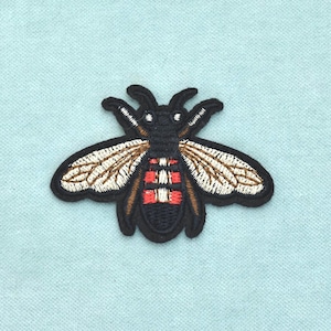 Op de afbeelding: Een geborduurde patch in de vorm van een bij. Het insect heeft een zwart lichaam met witte en rode horizontale strepen op het achterlijf, en vleugels gedetailleerd met beige en wit garen. Het ontwerp is afgewerkt met een zwarte rand.
