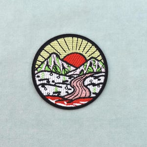 Può includere: Toppa ricamata circolare raffigurante un paesaggio montano con un sole rosso, montagne verdi e un fiume rosa. La toppa ha un bordo nero ed è ambientata su uno sfondo azzurro chiaro. Il design evoca la natura e l'avventura.