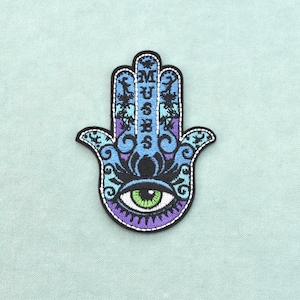 Puede incluir: Un parche bordado de mano de Hamsa con un ojo verde central, intrincados patrones decorativos en negro, azul claro y morado, y la palabra MUSES escrita verticalmente en letras mayúsculas negras en la sección del dedo medio.