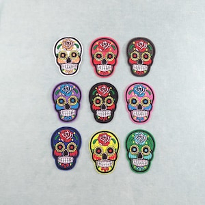 Peut inclure: Neuf écussons de crâne colorés avec des motifs floraux. Chaque écusson représente un crâne avec des orbites, des dents et une rose sur le front. Les écussons sont de différentes couleurs, dont le rouge, le jaune, le violet et le vert.