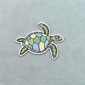 Può includere: Toppa ricamata con un disegno di tartaruga marina. Il guscio della tartaruga è un mosaico di forme geometriche verdi, gialle e blu. La toppa ha un bordo bianco su uno sfondo grigio chiaro.