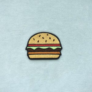 Op de afbeelding: Geborduurde patch van een hamburger met een sesambroodje, rode tomaat, groene sla en een bruine patty. De patch heeft een zwarte rand en is geplaatst tegen een lichtblauwe achtergrond.
