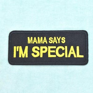 Può includere: Una toppa rettangolare nera ricamata con un bordo cucito. Il testo MAMA SAYS I'M SPECIAL è ricamato in lettere maiuscole giallo brillante al centro della toppa, con la parola SPECIAL che appare in un carattere di dimensioni maggiori.