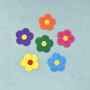 Op de afbeelding: Een set van zes geborduurde bloemapplicaties in diverse kleuren, waaronder oranje, paars, groen, roze, geel en blauw. Elke bloem heeft vijf afgeronde bloemblaadjes en een cirkelvormig hart, met een contrasterende kleur garen voor het detail.
