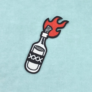 Puede incluir: Un parche bordado que muestra una botella de vidrio blanca con una etiqueta negra marcada con tres símbolos X blancos. La botella tiene un contorno negro y una llama roja brillante que emerge de la parte superior, diseño de cóctel Molotov.