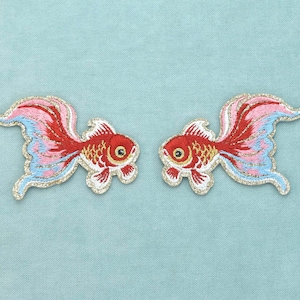 Op de afbeelding: Twee geborduurde patches in de vorm van goudvissen, elk met een rood lichaam met gouden schubben, een witte buik en vloeiende vinnen in tinten rood, roze en lichtblauw, allemaal omlijnd met een glinsterende gouden metallic draad.